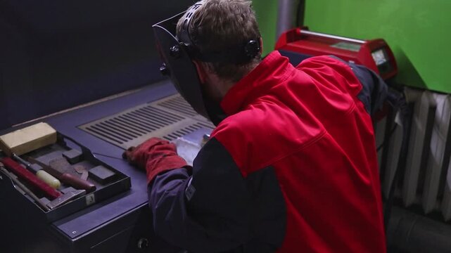 A student cleans a metal part with a metal brush after welding in a training workshop, preparing the seam for further processing and control.