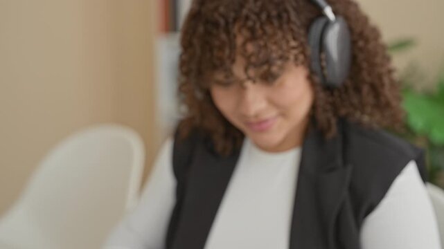 Woman wearing headphones writing notes at a desk in a building office setting with chair and plant visible; focused productivity.