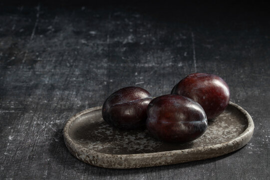 Minimalist dark food photography of ripe plums on an oblong ceramic tray on a distressed surface