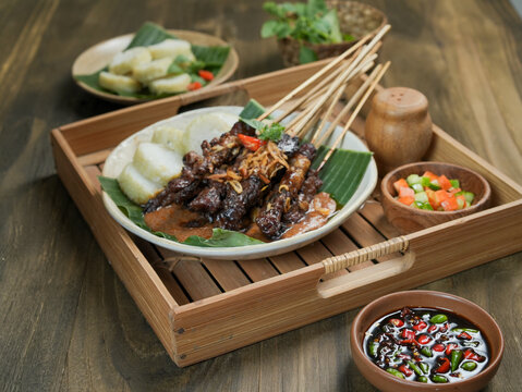 A hand picks grilled chicken satay with rice cakes and peanut sauce from a rustic wooden platter in Bandung Indonesia highlighting a traditional Indonesian dining experience.