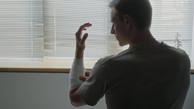 Caucasian young adult man raising bandaged arm, flexing fingers and rotating wrist to test mobility after injury. Video suitable for medical rehabilitation and health insurance ads