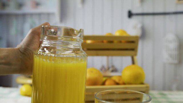 Person's hand pouring pitcher of juice into glass with surrounding oranges - slow motion closeup shot