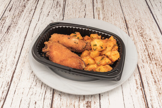 Breaded flamenquines paired with seasoned potato chunks in a black takeout tray on a whitewashed table.