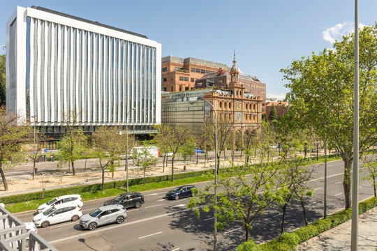 Traffic flows past modern office tower and historic tiled clock tower building on busy Madrid avenue