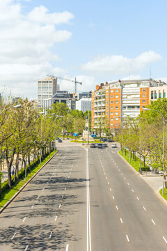 Wide multi-lane Madrid boulevard leading to statue roundabout, flanked by orange-brick apartment buildings and trees