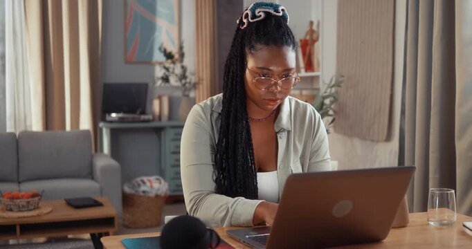 Young Black woman reads good news on her laptop at a desk in the living room and reacts with surprise and joy while celebrating her success at home.
