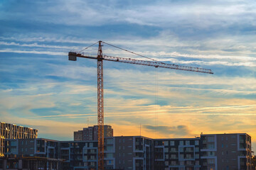 Fototapeta premium Tower Crane Above Modern Residential Buildings at colorful Sunset. Tower construction crane rising above modern residential apartment buildings. Urban development, housing construction, real estate