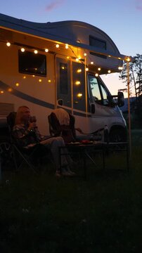 Blonde woman sitting in a folding chair enjoying a relaxing evening drink outside her motorhome, which is decorated with warm string lights under the awning during a scenic summer vacation