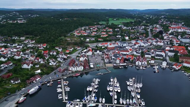 Aerial view of the coastal town of Lillesand with its marina filled with boats, white wooden houses with red roofs, and surrounding green hills in Lillesand, Agder, Norway.