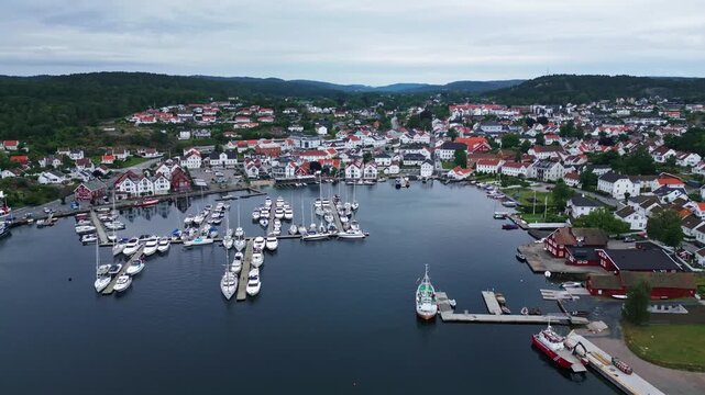 Aerial view of the picturesque harbor of Lillesand with numerous moored boats, white wooden houses, and lush green hills in the background in Lillesand, Agder, Norway.