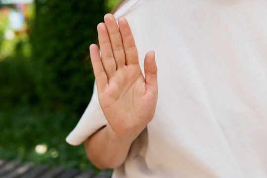 A person is showing a sign in sign language with their hand raised. The background includes greenery and soft light from the sun