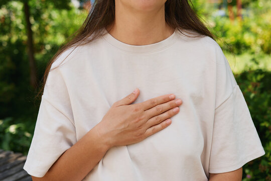 A person uses sign language with one hand on their chest while standing in a park. Trees and greenery are visible in the background