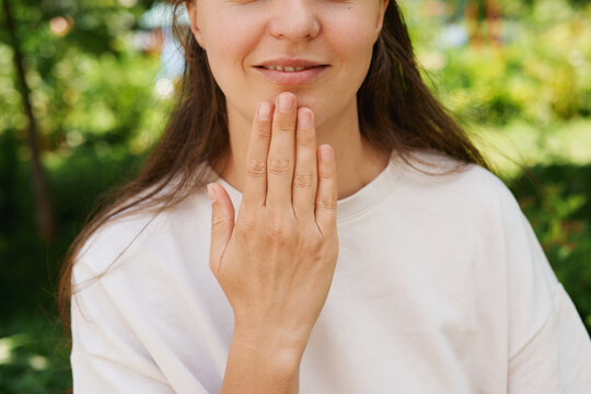 A woman stands in a park with green plants around her. She makes a sign language gesture with her hand near her face, focused and engaged