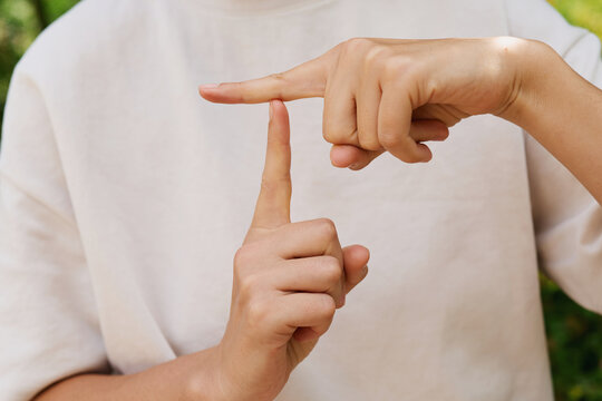Two hands are making signs in a green outdoor area. The fingers are positioned to communicate in sign language. It is daytime and the sun is shining