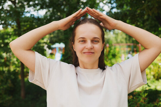 A woman is making a sign with her hands above her head in a park. She appears focused and engaged. The sun is shining and the trees are bright behind her