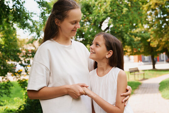 A mother and her daughter are smiling at each other as they use sign language to communicate. They are in a park surrounded by trees and grass