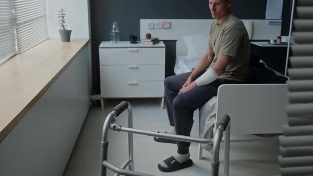 Tilt down shot of young adult Caucasian man with bandaged arm sitting on hospital bed near walker, looking out window. Video for healthcare, rehabilitation, and physical therapy promotions