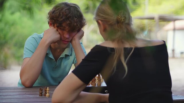 A teenage boy with curly hair concentrates on a chess game, sitting opposite his mother at a wooden table in an outdoor park