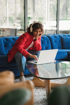 Vertical shot of curly haired latina woman leaning toward table, using laptop, types wearing headphones, smiling gently. Young hispanic freelancer works remotely from couch at home. Copy space