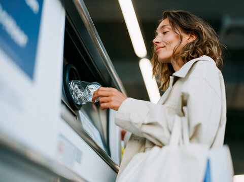 Smiling young woman returning a plastic bottle at a reverse vending machine in a supermarket, deposit return scheme and circular economy concept.