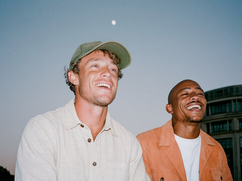 Two multiethnic male friends laughing together on a city rooftop at dusk, candid flash photo with authentic joy and urban skyline background.