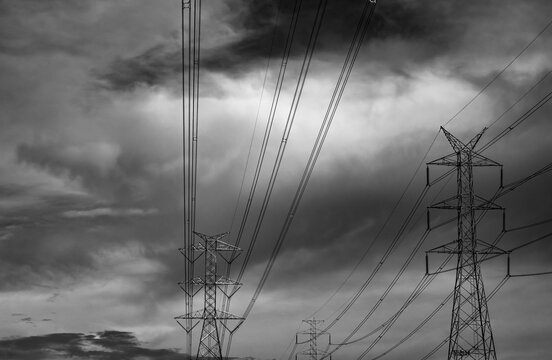 High voltage electricity transmission towers beneath dramatic storm clouds. Power grid infrastructure, energy security, electrical transmission network and climate resilience during severe weather.