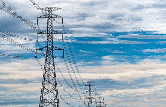 High voltage electricity transmission towers and power lines beneath a blue cloudy sky. Modern electrical infrastructure, utility grid reliability, energy distribution and sustainable power networks.