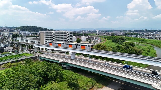 Aerial view of the Tama Toshi Monorail crossing the Asakawa River with lush greenery, residential buildings, and a road in Hino, Tokyo, Japan.