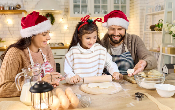Christmas happy family baking cookies together at home. Parents help child daughter roll dough in a cozy kitchen, wearing Santa hats amid lights and festive tree. Warm winter holidays