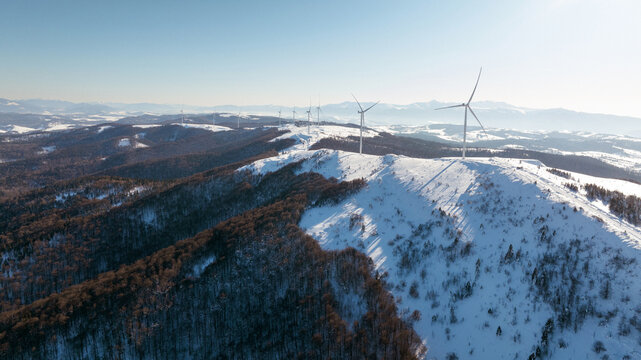 Wind turbines stand tall on a snow-covered mountain ridge under a clear blue sky
