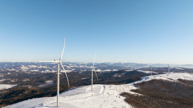 Wind turbines stand tall on a snow-covered mountain range under a clear blue sky