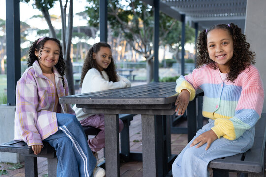 Three female youths sitting at dark picnic table in park pavilion, wearing colorful clothes