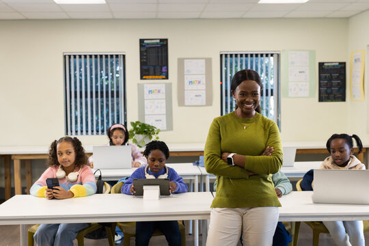 African American teacher standing front of classroom in sweater and smartwatch, children on laptops