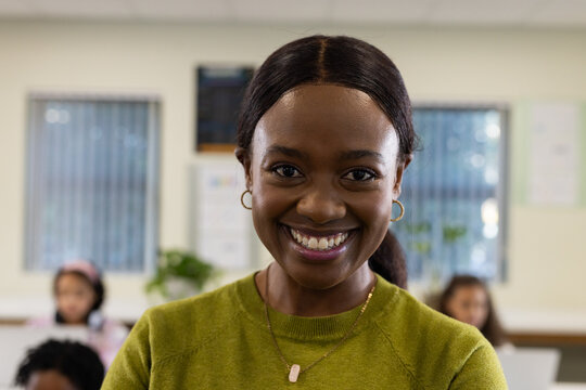 African American female teacher smiling in class in green sweater, gold pendant, students using PCs