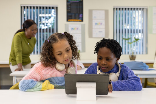 Female children sitting side-by-side at classroom table using tablet, wearing headphones, sweaters