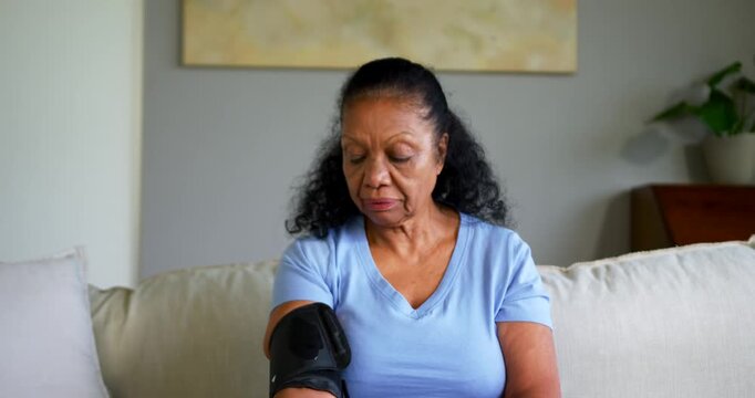 Senior African American woman securing cuff, monitoring tubing, checking blood pressure on couch