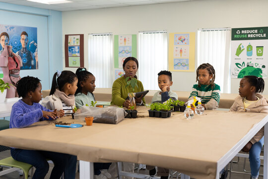 African American teacher with six youths guiding planting at classroom table with tablet, seedlings