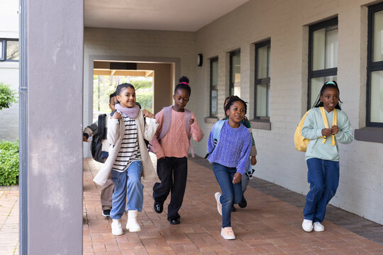 African American classmates hurrying along brick walkway beside school, carrying backpacks, scarves