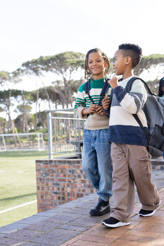 Two African American boys in striped sweaters walking on brick ledge by soccer field with backpacks