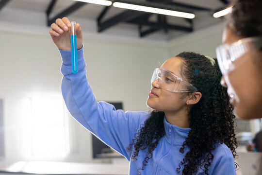 Two students with female youth wearing safety goggles and blue zip-up inspecting test tube in lab
