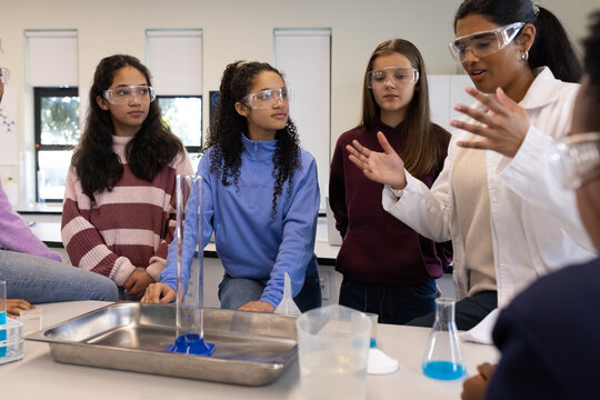 Diverse female students watching Indian instructor in lab coat at lab bench with graduated cylinder