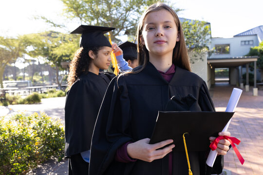 Diverse teen grads posing on sunny campus in gowns holding red-ribbon diploma and yellow-tassel cap