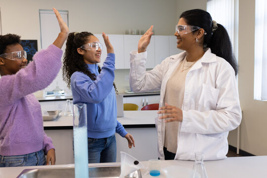 African American teen girls  and  Indian teacher high fiving in lab by blue cylinder  and  labcoat,g
