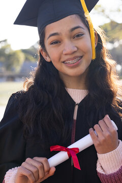 African American teen smiling holding diploma red ribbon wearing cap gold tassel gown sweater park