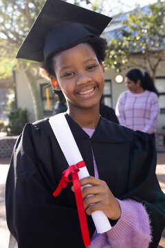 African American teen with Indian guardian smiling, holding diploma on campus, wearing black gown