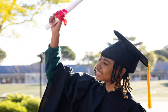 African American teen raising red-ribbon diploma in yellow-tassel cap,gown green cuff, copy space