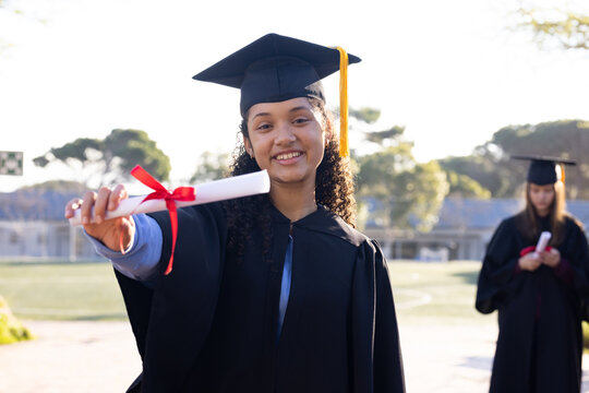 Diverse youth female graduates smiling and extending diploma scroll on school field in black gowns