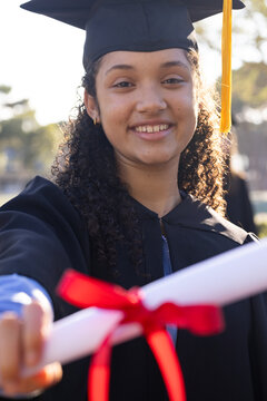 African American teen grad offering diploma with red ribbon on sunny campus in cap and gown