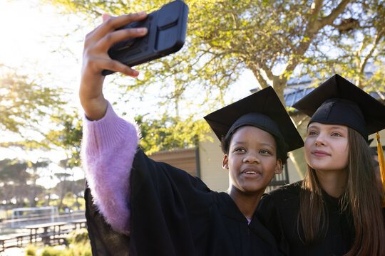Diverse female graduates holding smartphone in case posing for selfie on campus in caps and gowns