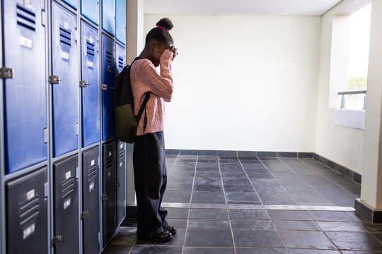 African child standing, hands covering face in school hall at lockers, in pink sweater, copy space